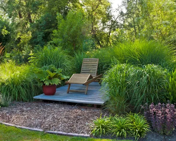 Entspannende Garten-Ideen: Eine Holzliege auf einer kleinen Terrasse inmitten üppiger Gräser und Pflanzen.