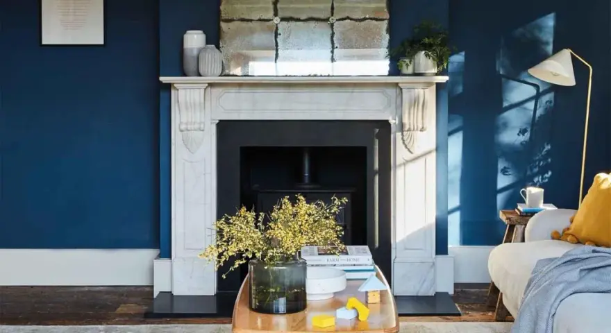 Ein Wohnzimmer mit tiefblauen Wänden. Ein Marmorkamin mit Spiegel darüber. Ein Holztisch mit Blumen und Spielzeug. Passt beige zu blau? Ja, die gelben Akzente und der beigefarbene Teppich zeigen es.