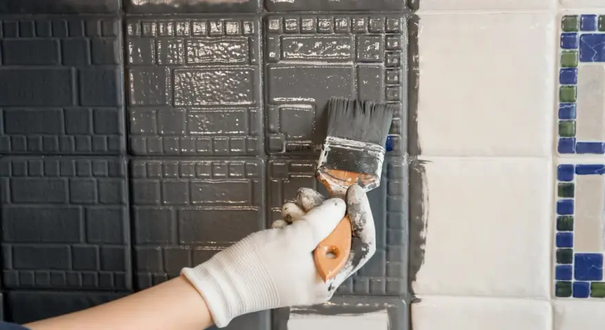 Hand mit Handschuh streicht mit Pinsel graue Farbe auf Fliesen. Ein Teil der Wand ist bereits grau, der andere noch weiß.