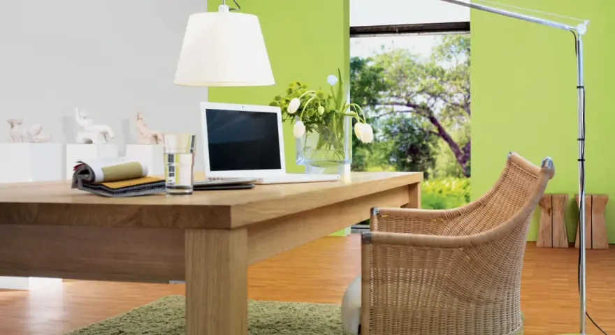Ein Schreibtisch aus Nussbaumholz mit Laptop und Stoffmustern. Grüne Wand und Rattanstuhl. Welche Farbe passt zu Nussbaum? Grün und Beige.