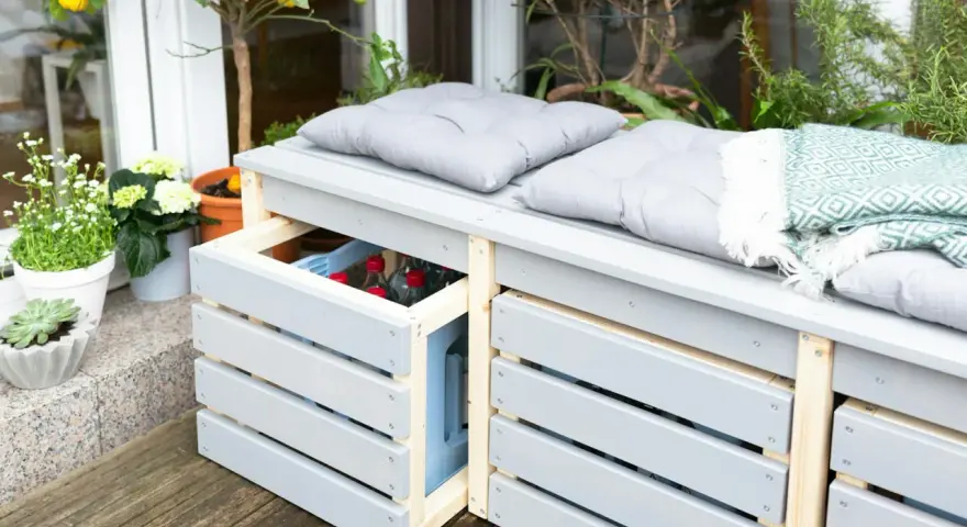Ordnung im Garten: Eine graue Gartenbank mit Stauraum für Getränkekisten und Kissen. Pflanzen und eine Decke sorgen für Gemütlichkeit.