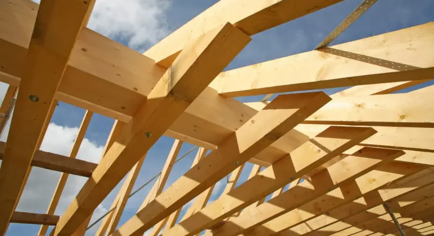 Holzkonstruktion eines Dachstuhls im Bau, mit sichtbaren Balken und Streben. Das Sprengwerk des Dachstuhls wird durch das Sonnenlicht beleuchtet.