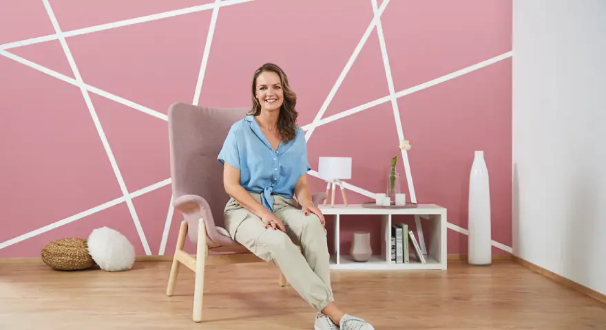 Frau sitzt vor einer rosa Wand mit weißem geometrischem Muster. Tolle Ideen zum Wand streichen und Muster abkleben!