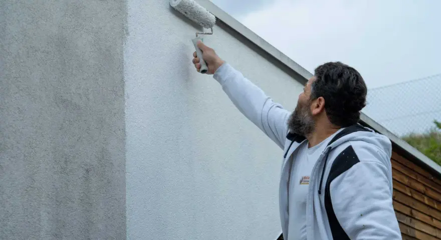 Ein Mann mit Bart streicht mit einer Malerrolle eine Hauswand. Die Wand zeigt horizontale Streifen, da sie gerade neu gestrichen wird.