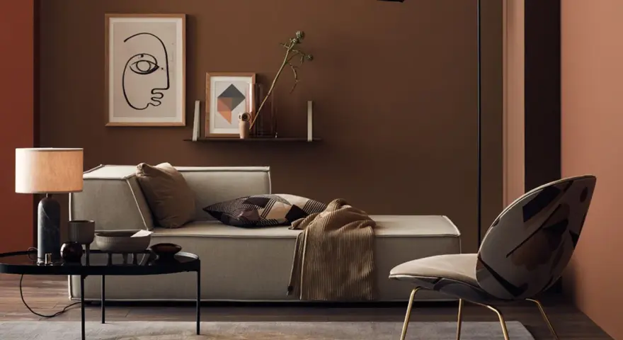Gemütliches Wohnzimmer mit Sofa, Sessel und Tisch. Die Wände sind in warmen Brauntönen gehalten, die sich mit den Textilien und Dekorationen **braun mischen**.