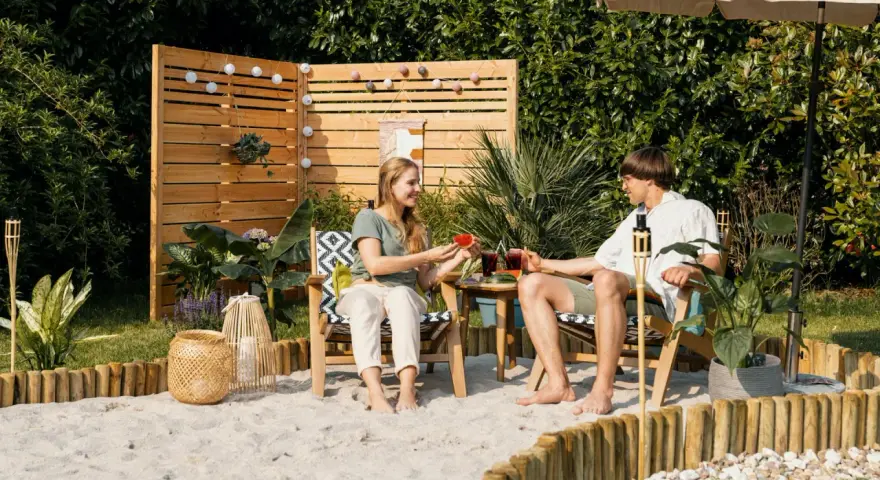 Ein Paar genießt den Sommer mit Wassermelone und Drinks im Garten. Sand, Holz und Pflanzen schaffen ein echtes Strandfeeling im Garten.