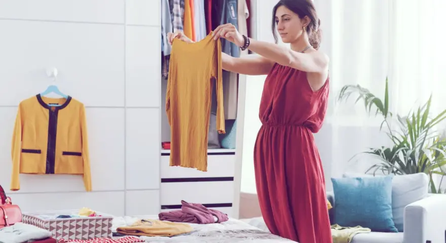 Frau sortiert Kleidung nach der Marie Kondo Methode. Ein gelbes Oberteil wird in die Hand genommen, daneben ein gelber Blazer und ein Korb mit gefalteten Textilien.