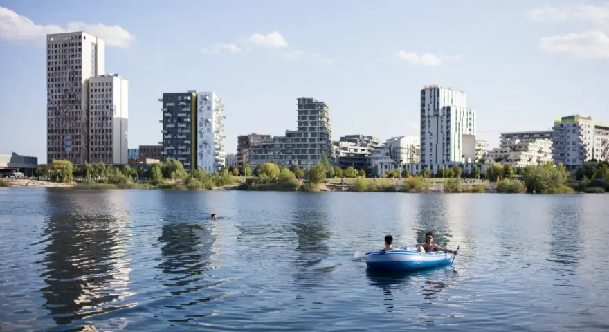 Zwei Männer treiben in einem Schlauchboot auf dem See der Seestadt Aspern. Im Hintergrund ragt die moderne Architektur der Stadt auf.