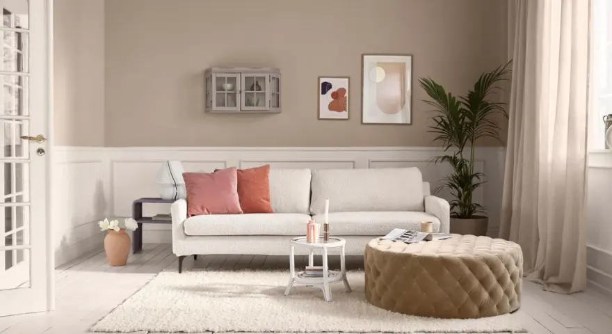 Gemütliches Wohnzimmer mit cremefarbener Couch, passenden Kissen und einem Hocker. Die ecru farbe kombinieren sich harmonisch mit den Wandfarben und Deko-Elementen.
