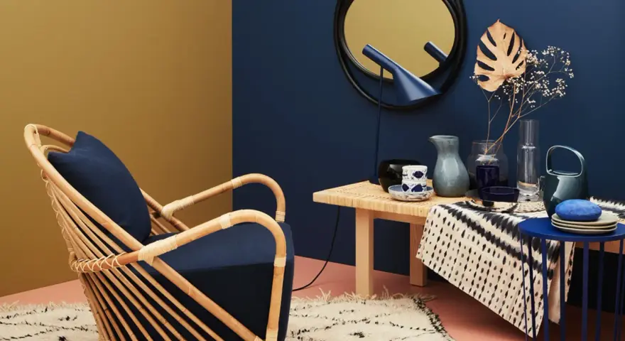 Ein Rattansessel mit dunkelblauem Kissen steht auf einem Teppich. Welche Farbe passt zu Gold? Das goldene Spiegelbild an der Wand und die gelbe Wandfarbe harmonieren mit dem tiefen Blau.