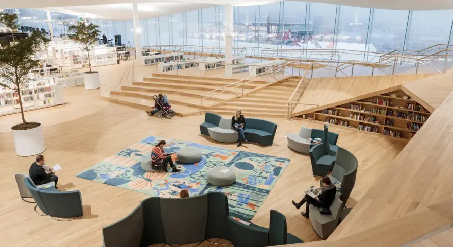 Helle, offene Bibliothek in Oodi Helsinki mit Menschen, die lesen und sich entspannen. Holzmöbel und große Fenster schaffen eine einladende Atmosphäre.