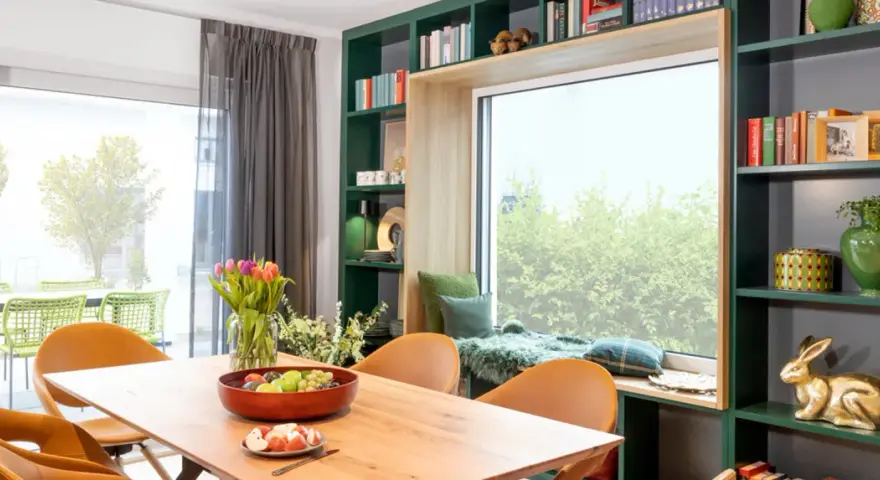 Gemütliche Leseecke mit Kissen und Decke am Fenster gestalten. Ein Holztisch mit Obstschale und Tulpen steht davor. Grüne Regale mit Büchern und Deko umrahmen das Fenster.
