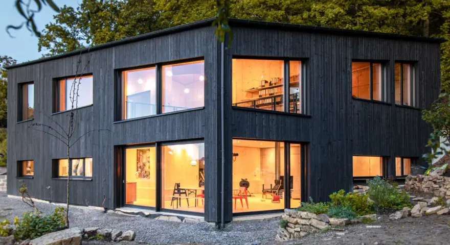 Modernes Haus mit Fassade im Shou Sugi Ban Stil. Große Fenster lassen warmes Licht herein.