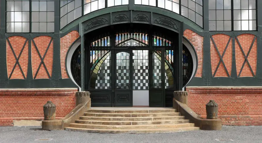 Eingang im artdeco stil mit Ziegelmauerwerk, geschwungenen Metallverzierungen und einem beeindruckenden Buntglasfenster.