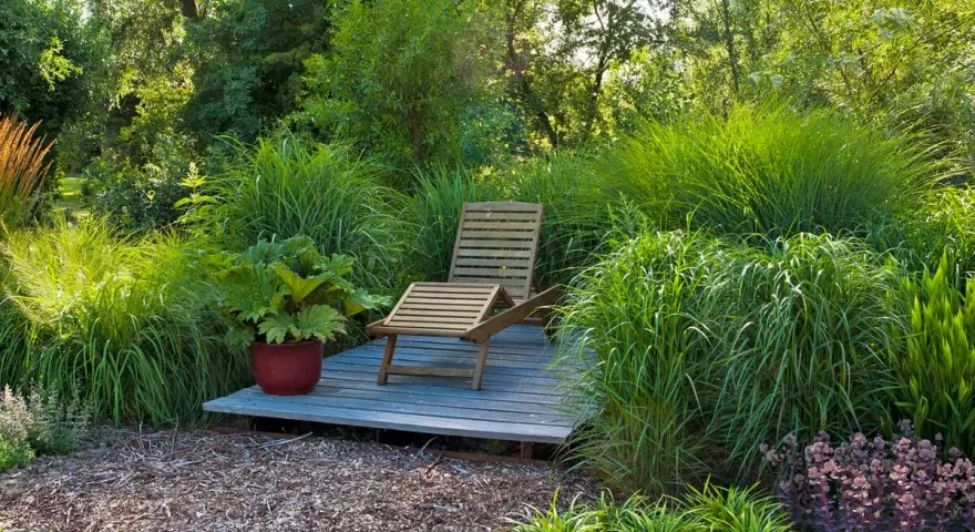 Entspannende Garten-Ideen: Eine Holzliege auf einer kleinen Terrasse inmitten üppiger Gräser und Pflanzen.