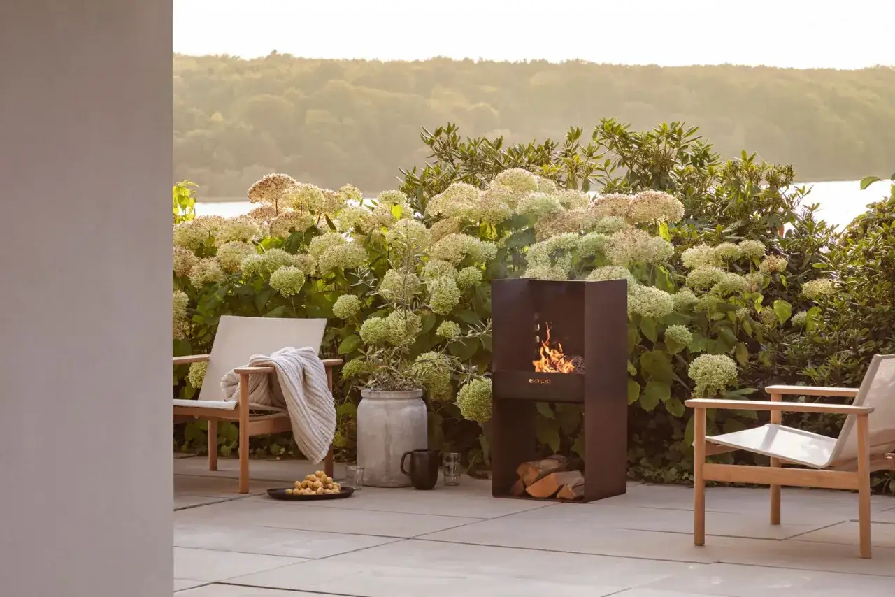 Gem&uuml;tliche Gartendeko Ideen: Ein moderner Feuers&auml;ule mit brennendem Holz, daneben zwei Sessel und Hortensien.