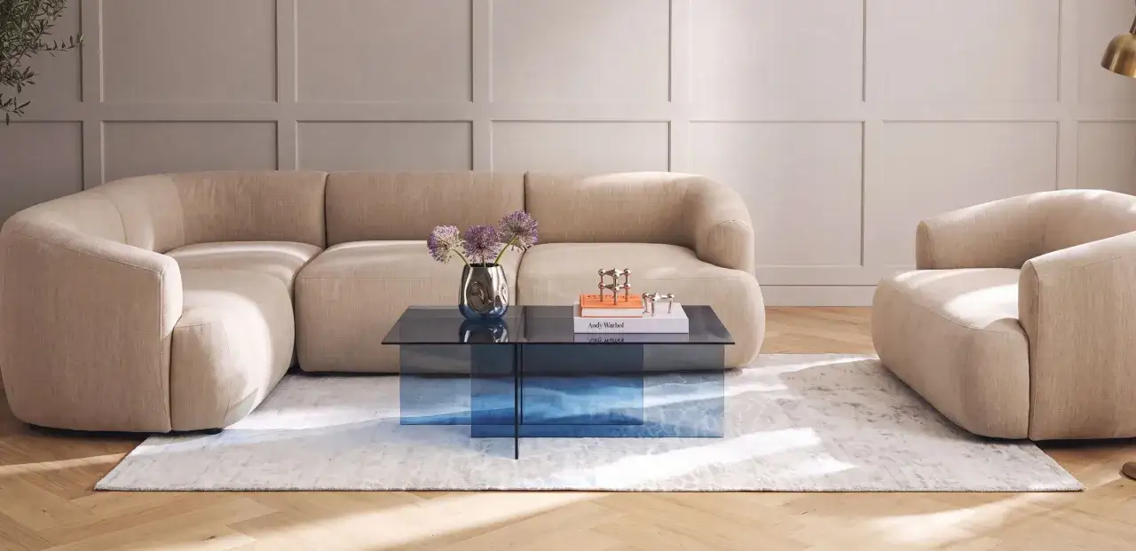 Beige Sofas und ein Sessel bilden eine gemütliche Ecke. Ein blauer Glastisch mit Blumen und Büchern ergänzt die Szene und zeigt, wie gut passt beige zu blau.