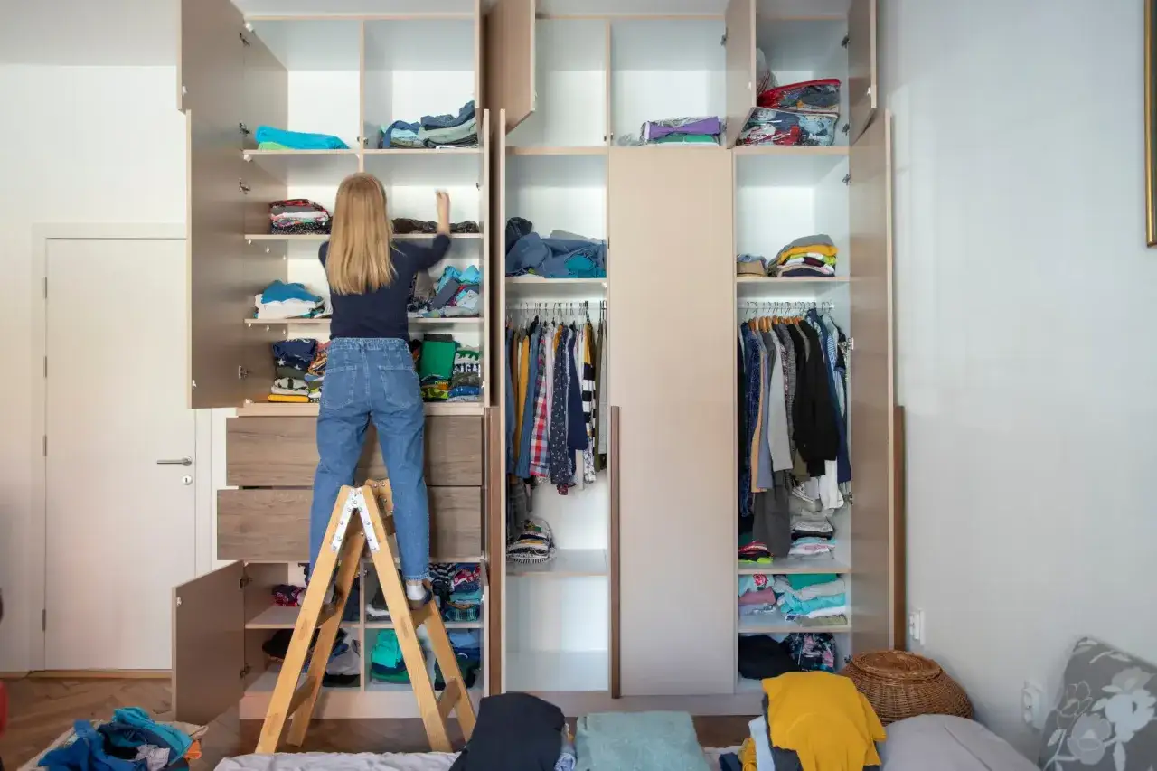 M&auml;dchen auf Trittleiter sortiert Kleidung im offenen Kleiderschrank. Gute ausmisten tipps helfen, Ordnung zu schaffen.