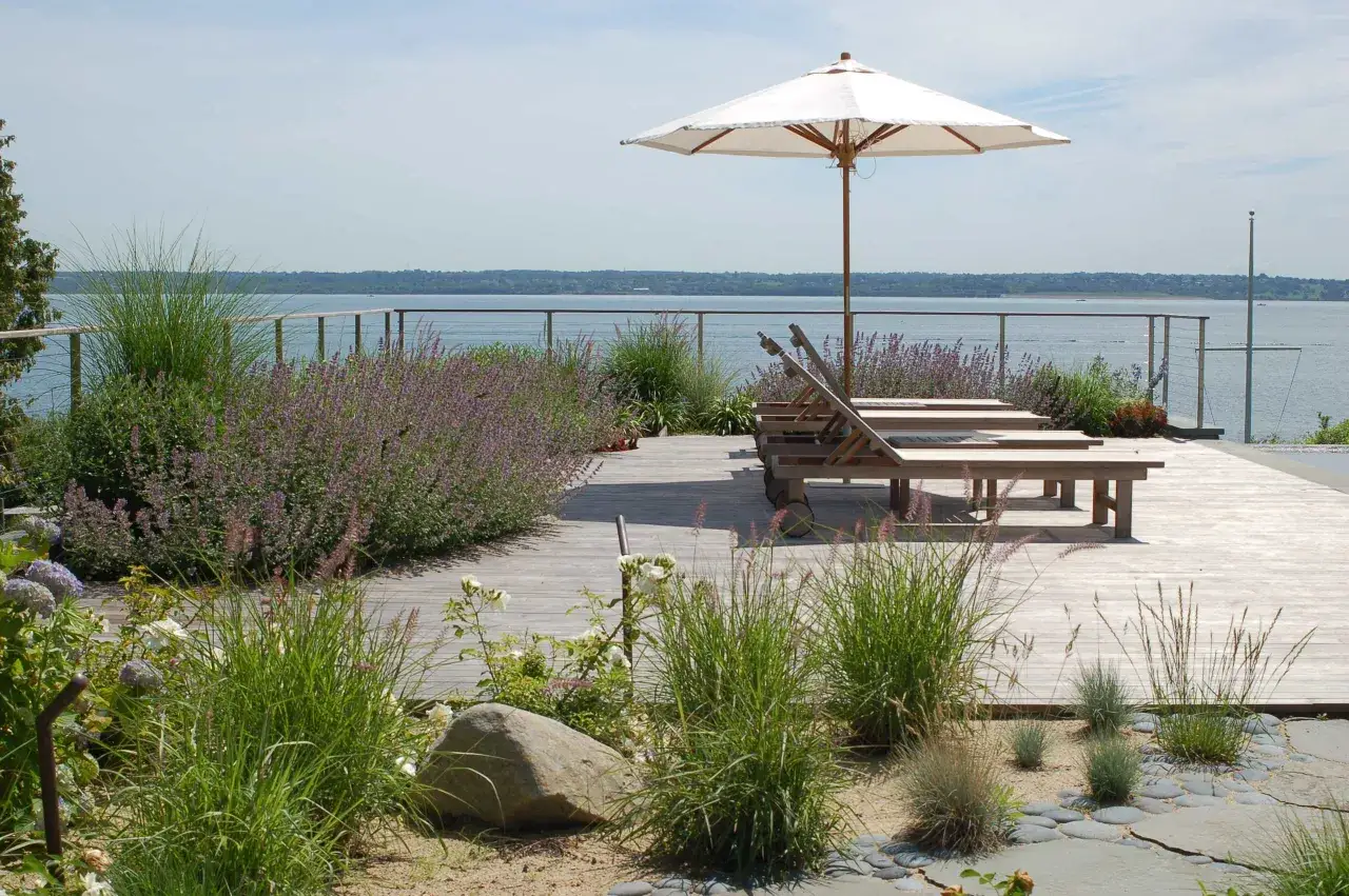 Holzdeck mit Liegest&uuml;hlen und Sonnenschirm, umgeben von Gr&auml;sern und Lavendel. Ein Hauch von Strandfeeling im Garten mit Blick aufs Wasser.