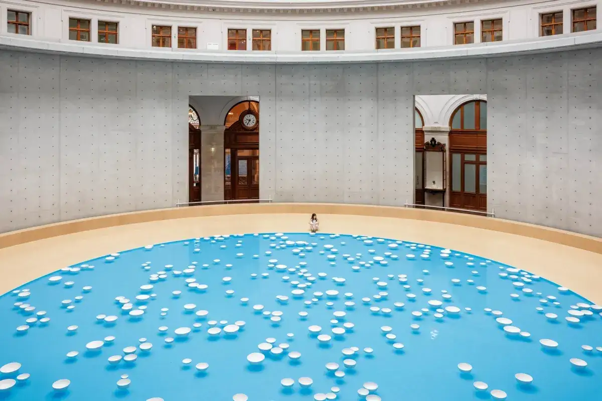Ein M&auml;dchen sitzt in der Mitte eines blauen Beckens mit vielen wei&szlig;en Scheiben, das an eine futuristische Bourse de commerce erinnert.