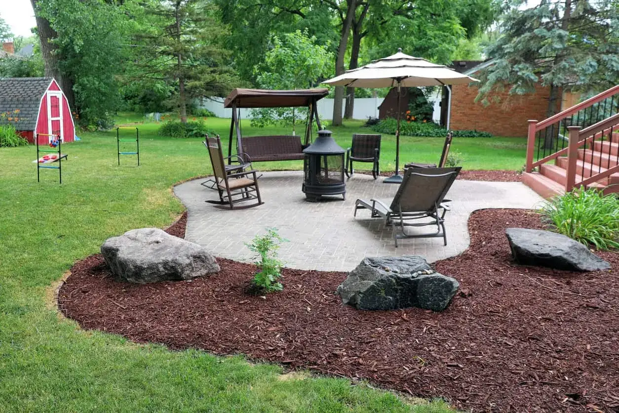 Rindenmulch garten ideen: Einladende Terrasse mit Schaukel, Feuerstelle und Liegest&uuml;hlen, umgeben von Rasen und dekorativem Rindenmulch.