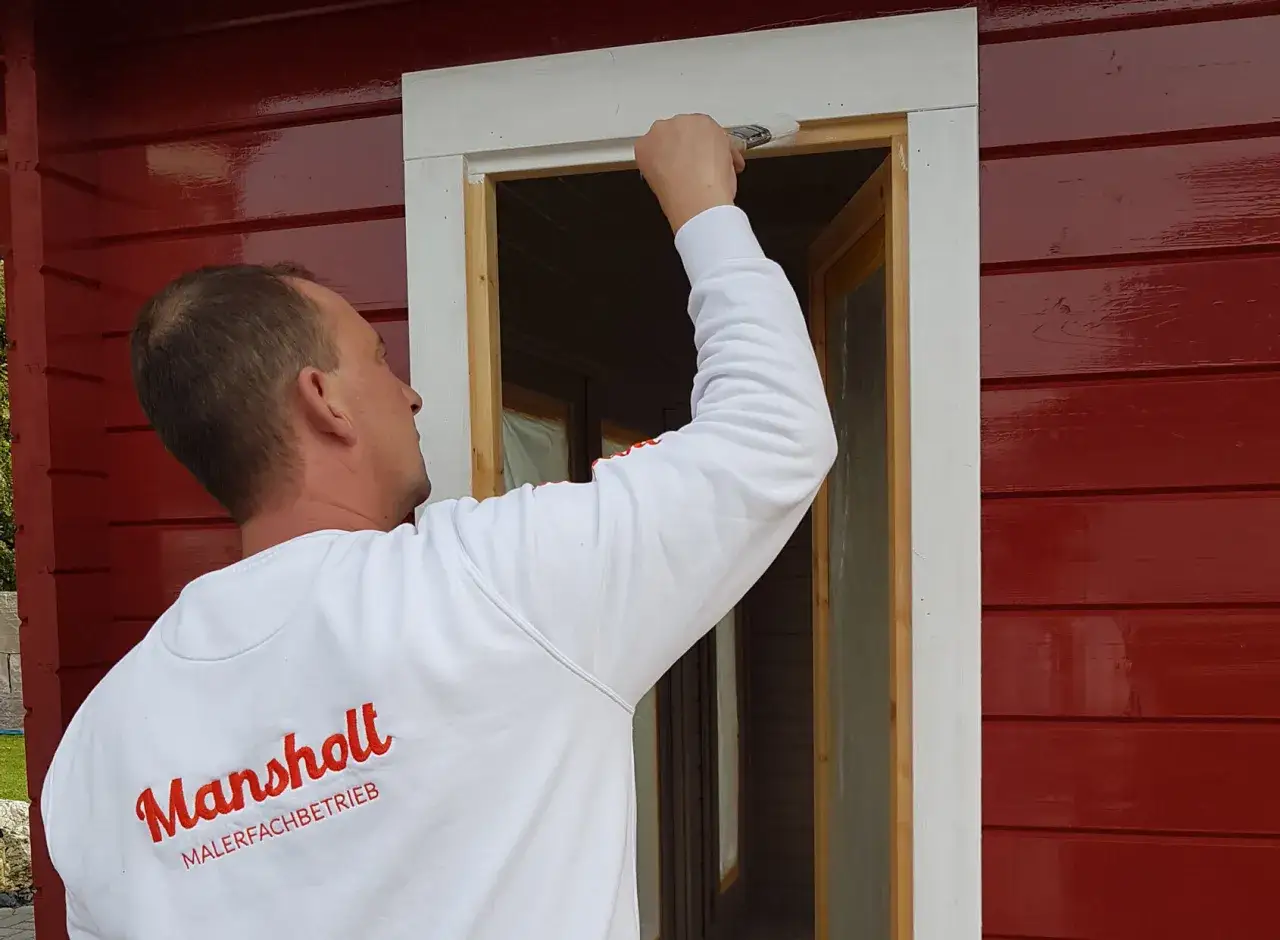 Maler streicht mit Pinsel die weiße Wandfarbe auf das Holz des Fensterrahmens.