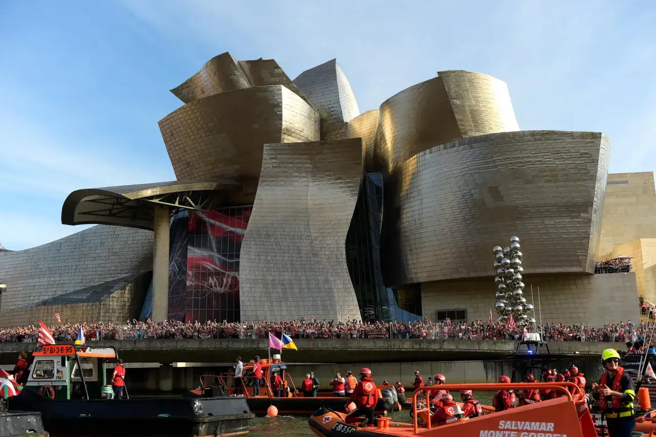 Frank Gehry Bauwerke: Menschen jubeln am Ufer des Guggenheim Museums Bilbao, w&auml;hrend Rettungsboote auf dem Wasser fahren.