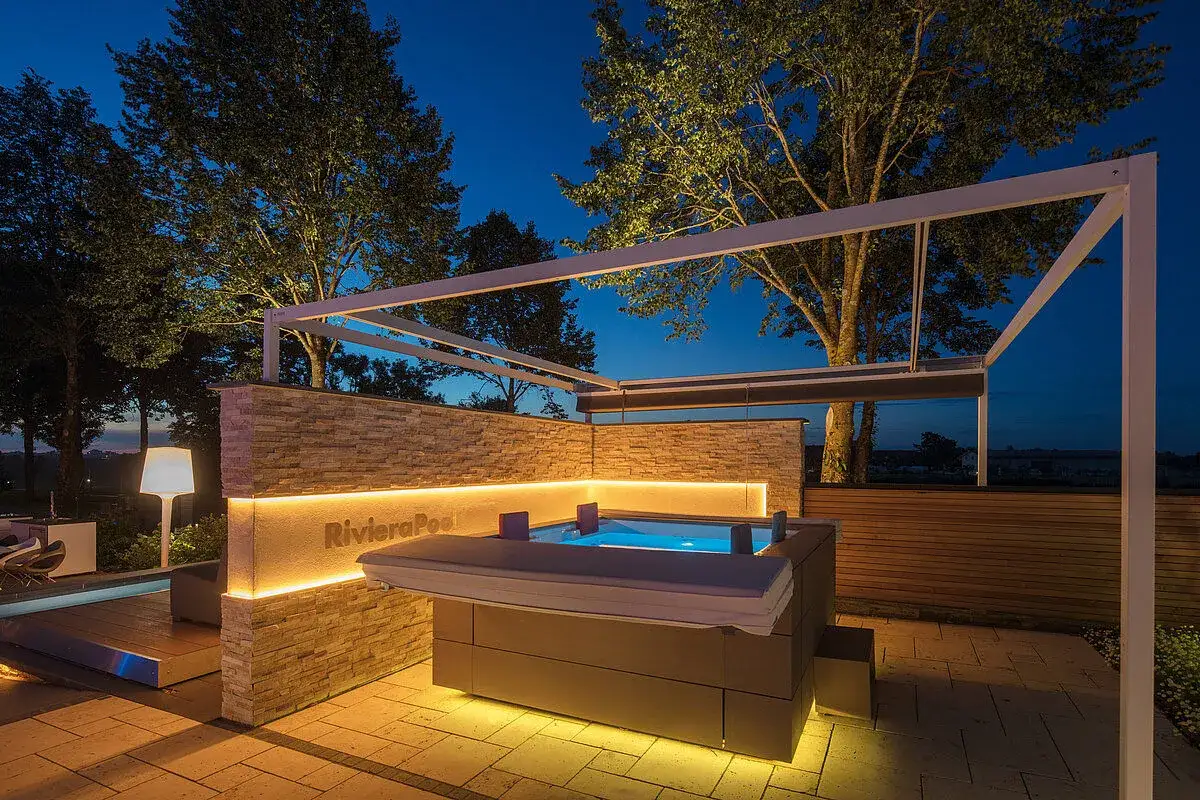 Ein luxuri&ouml;ses Jacuzzi mit beleuchteter Steinwand und Pergola im Abendlicht. Hier kann man sich fragen: was ist ein Jacuzzi?