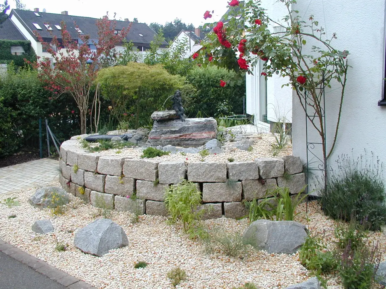 Moderner Hauseingang mit einem geschwungenen Steingarten und einem kleinen Wasserspiel. Ein Rosenbogen &uuml;berdacht den Eingangsbereich.