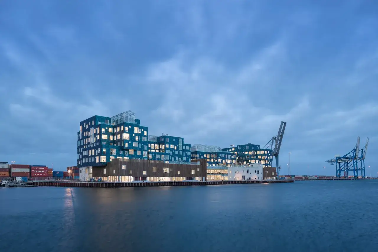 Moderne Architektur am Nordhavn Kopenhagen: Ein Gebäude mit blauen und hölzernen Fassaden spiegelt sich im Wasser, Container und Kräne im Hintergrund.