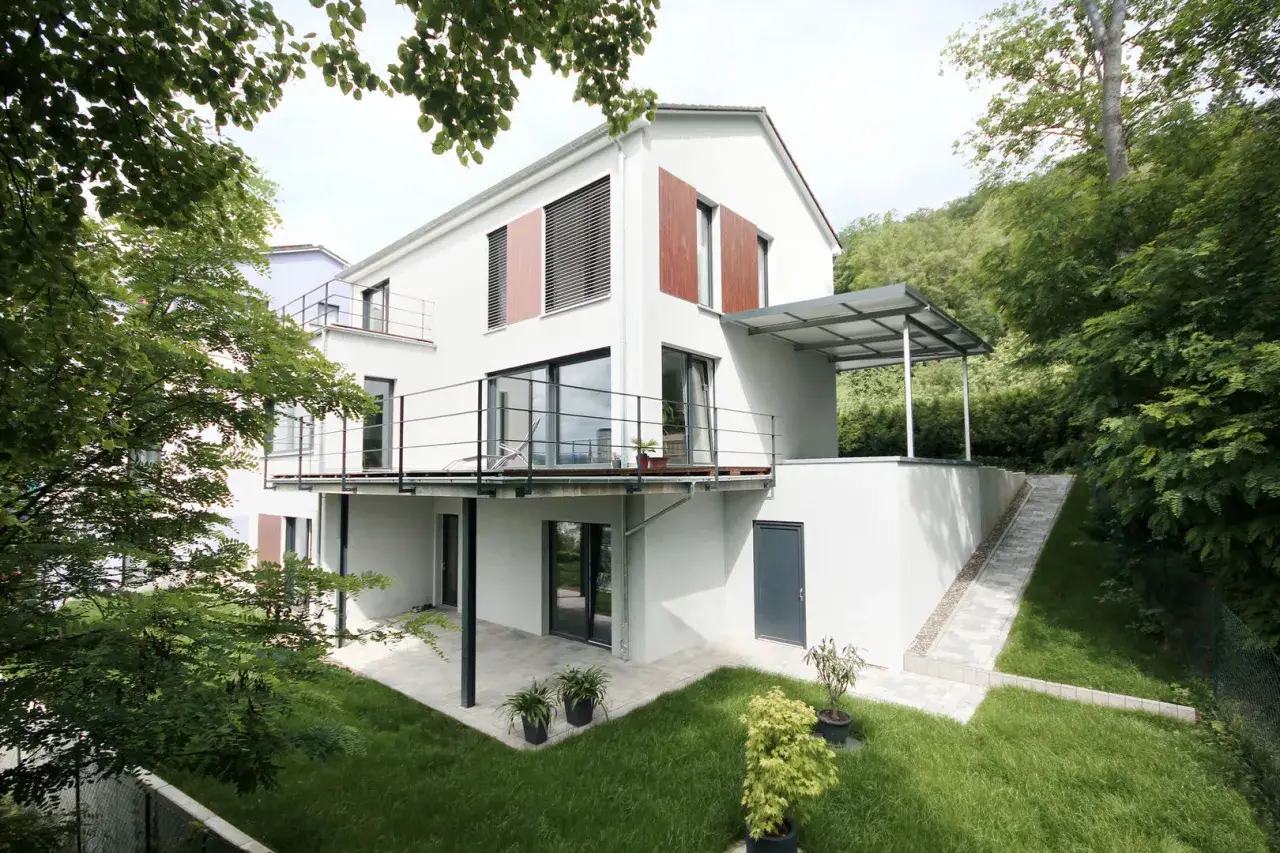 Modernes Haus mit Balkonen und einer &uuml;berdachten Terrasse, die als Veranda dient. Gr&uuml;ne Rasenfl&auml;chen und B&auml;ume umgeben das Geb&auml;ude.