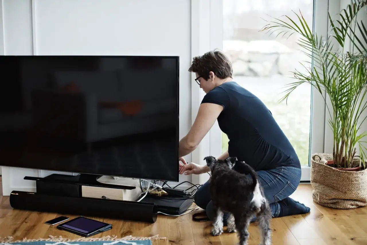 Frau mit Hund richtet Kabel am gro&szlig;en Fernseher im Raum.