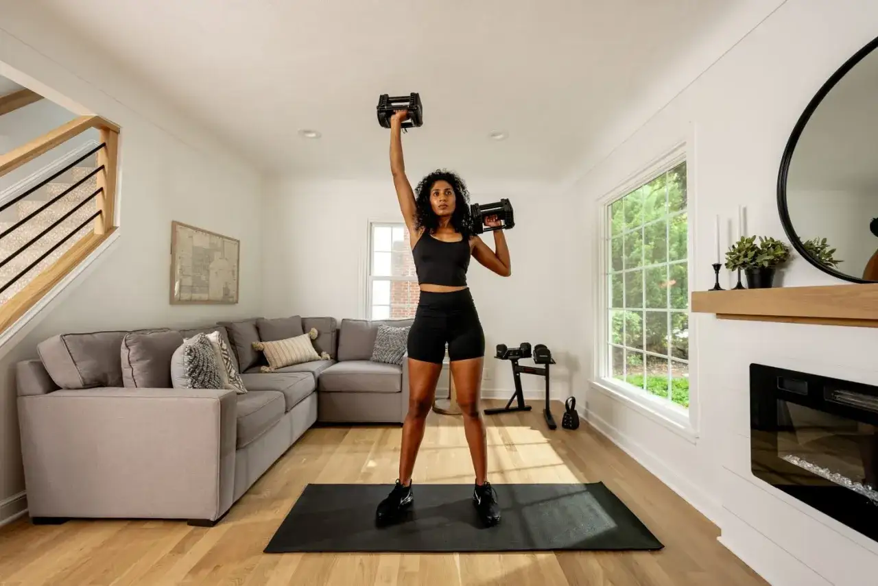Frau trainiert mit Hanteln in ihrem kleinen Fitnessraum zuhause. Ein Sofa und eine Treppe sind im Hintergrund zu sehen.