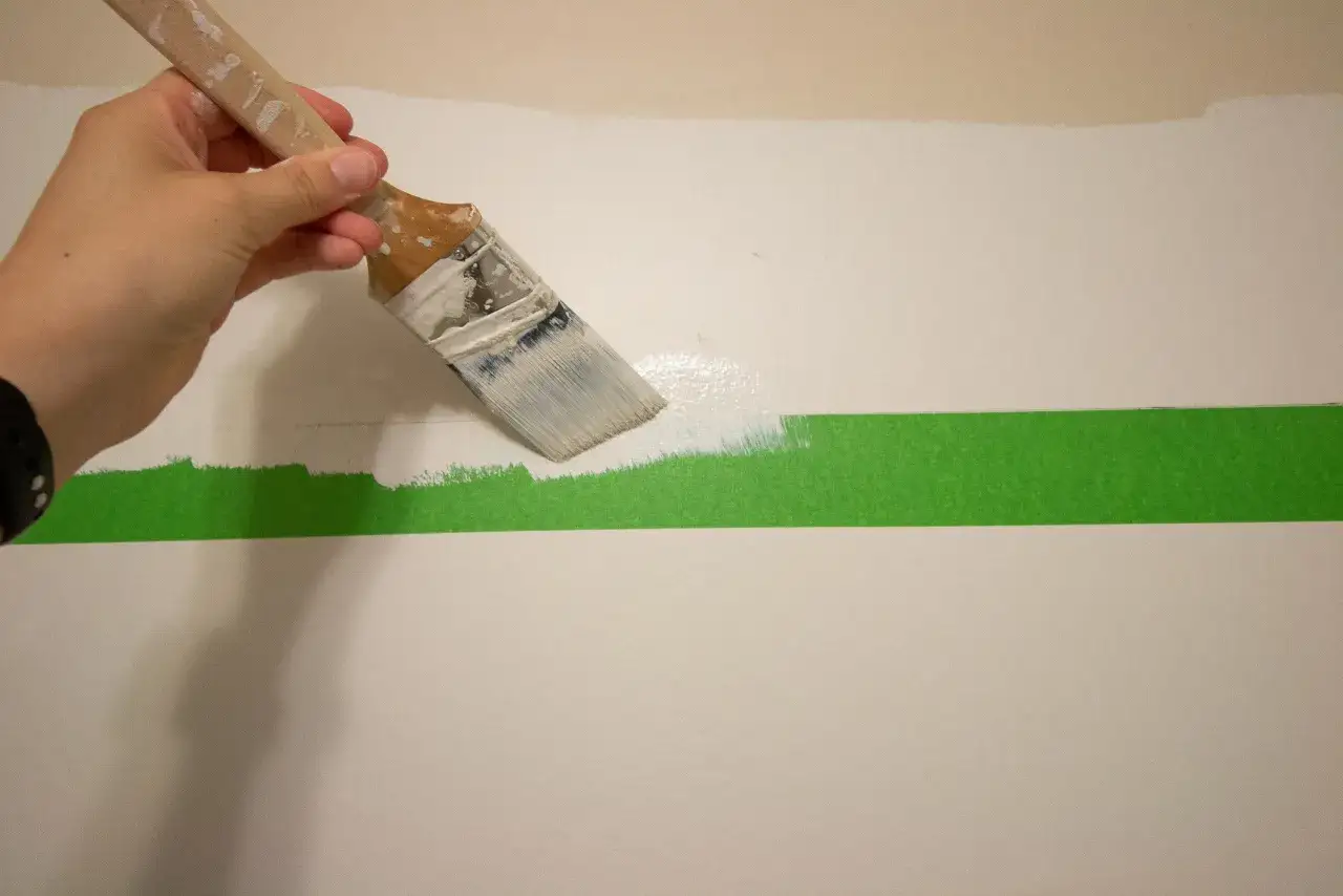 Hand streicht mit Pinsel wei&szlig;e Farbe &uuml;ber eine Wand, die mit gr&uuml;ner Klebefolie abgeklebt ist, um einen sauberen Rand zu schaffen.