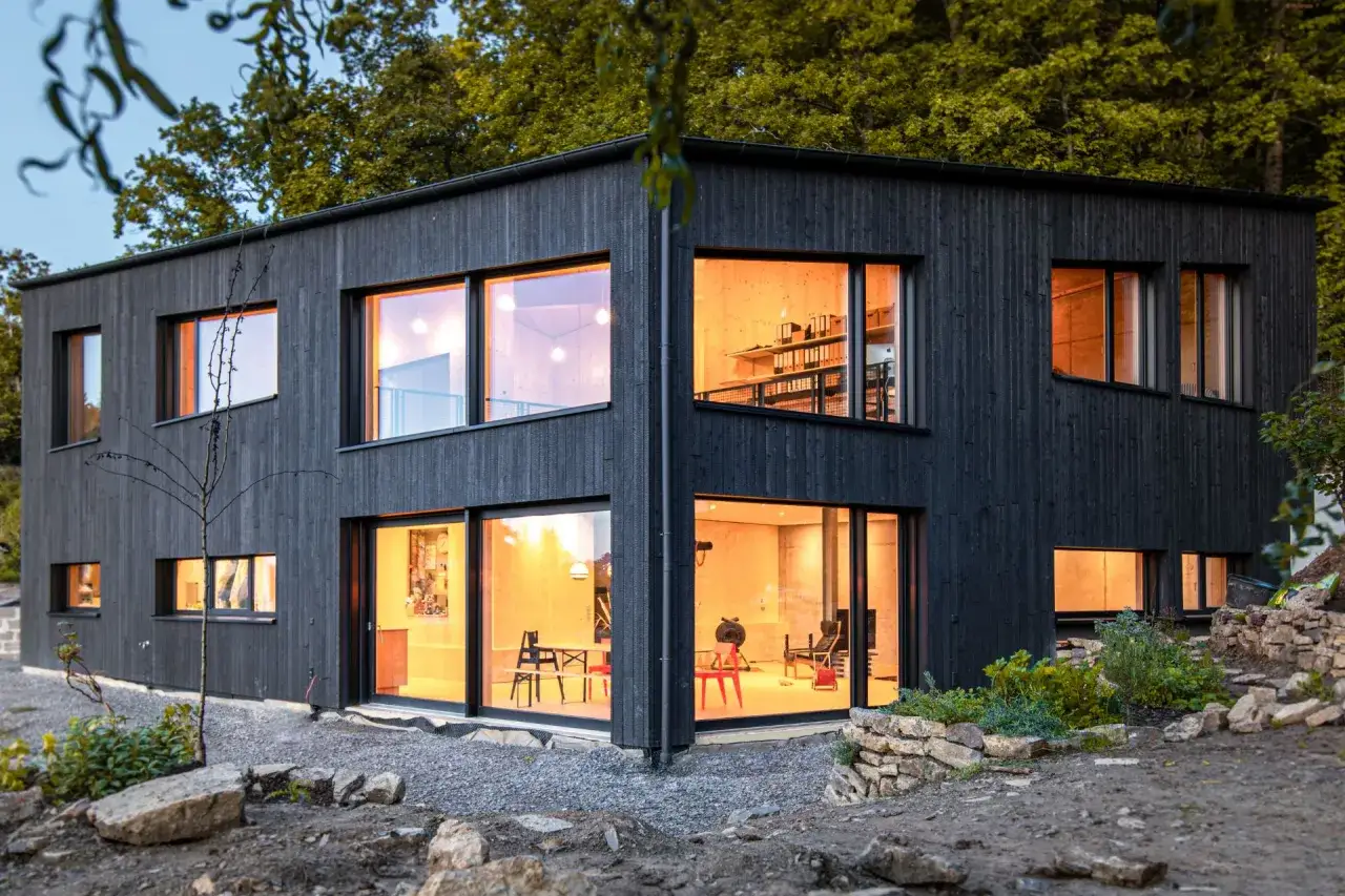 Modernes Haus mit Fassade im Shou Sugi Ban Stil. Gro&szlig;e Fenster lassen warmes Licht herein.