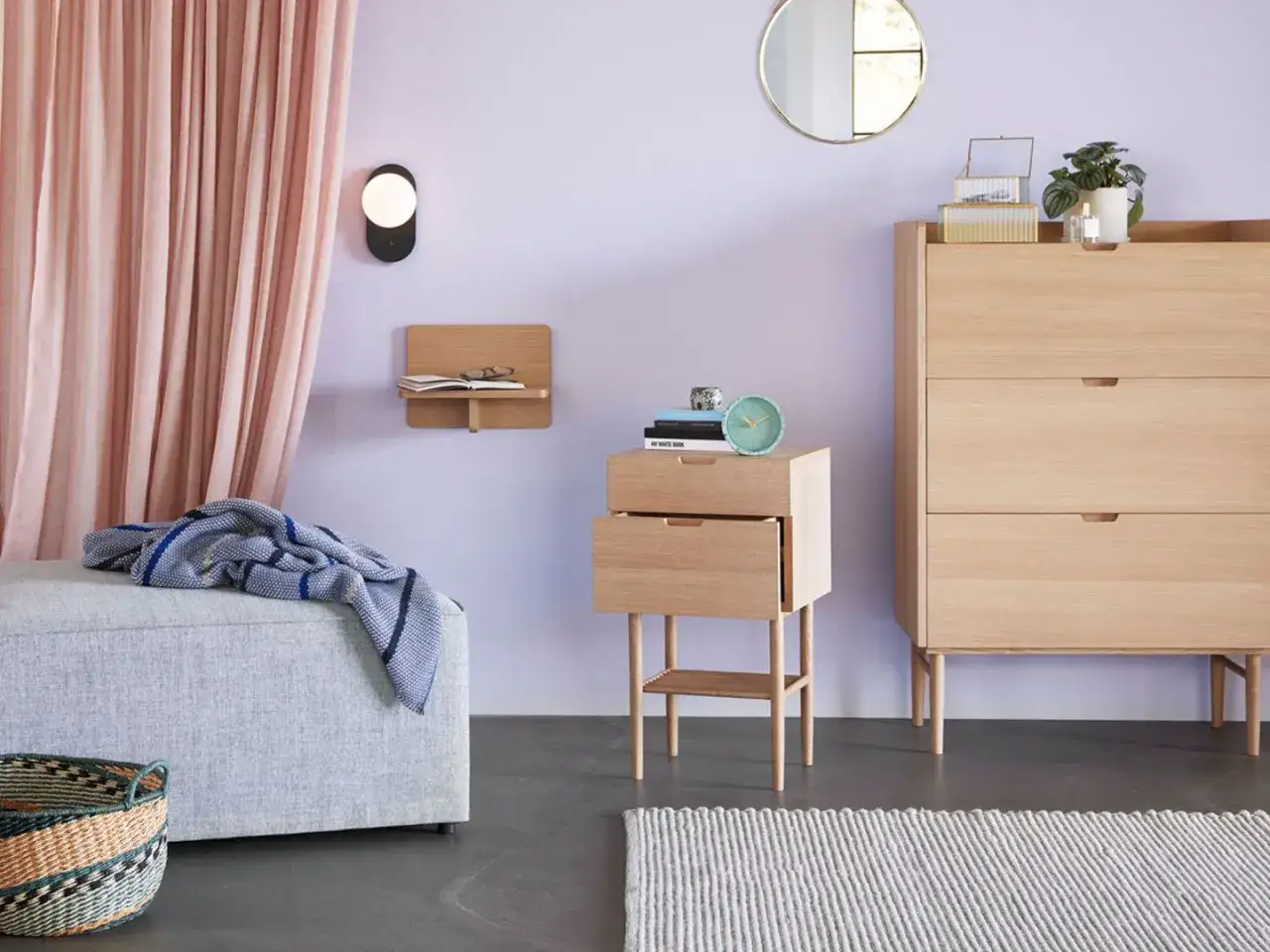 Ein Schlafzimmer mit hellen Holzm&ouml;beln und einer rosafarbenen Gardine. Die Wandfarbe ist ein sanftes Lila, das mit dem Licht zu spielen scheint. Ein kleiner Nachttisch mit B&uuml;chern und einem Wecker steht neben einem Bett. Ein Korb und eine Decke vervoll...