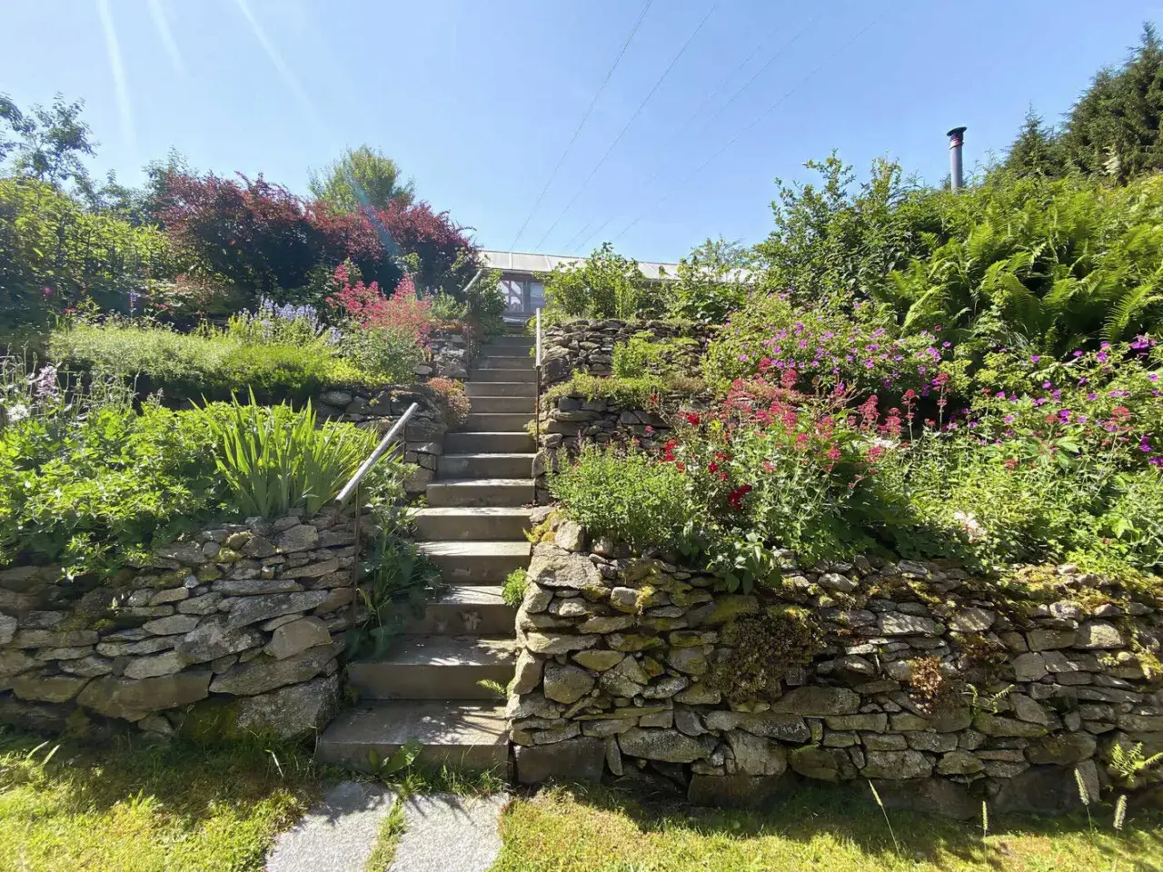 Blumen und Steine schm&uuml;cken die Gartengestaltung Hanglage. Eine Treppe f&uuml;hrt durch &uuml;ppige Beete zu einem Gew&auml;chshaus.