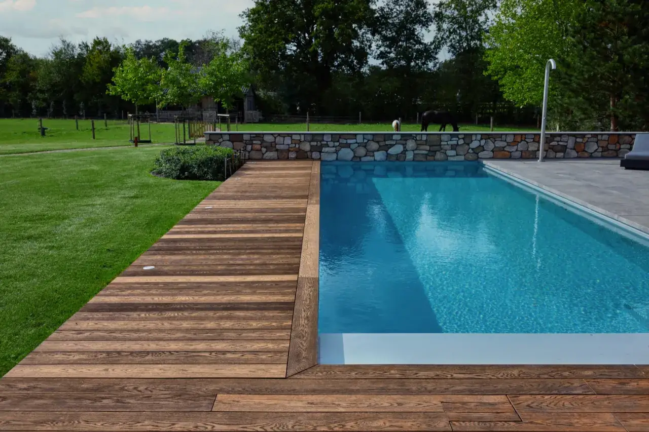 Ein moderner Pool mit Holzdeck und Steinmauer. Tolle Ideen Schwimmteich f&uuml;r ein naturnahes Gartendesign mit Pferden im Hintergrund.