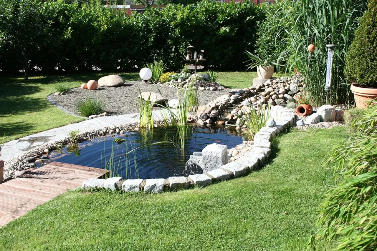 Ein idyllischer kleiner gartenteich ideen mit Holzsteg, Steinumrandung und Wasserfall. &Uuml;ppige Pflanzen und ein gepflegter Rasen umgeben das Wasser.