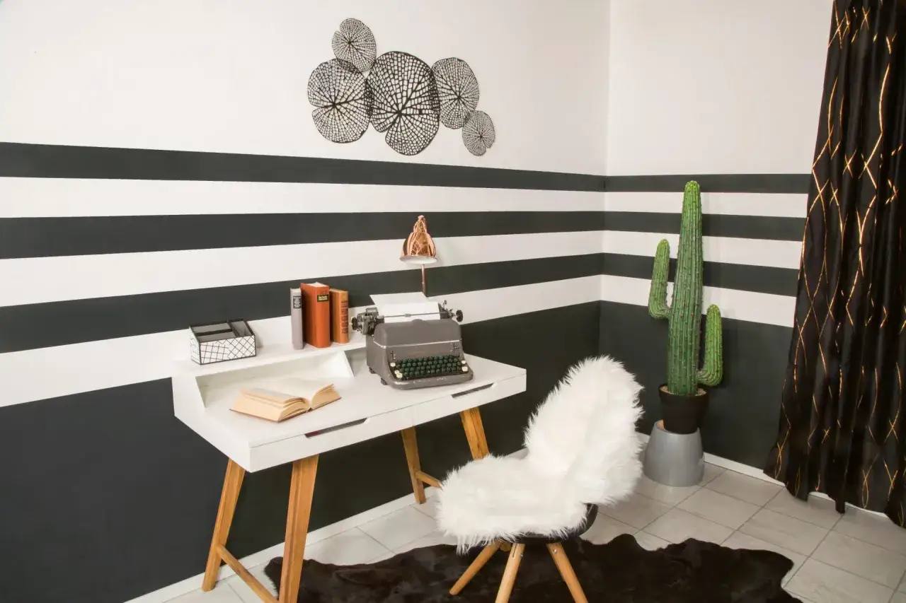 Ein Schreibtisch mit Schreibmaschine und Büchern steht vor einer Wand mit horizontalen Streifen. Ein flauschiger Stuhl und eine Kakteen-Dekoration vervollständigen das Bild.
