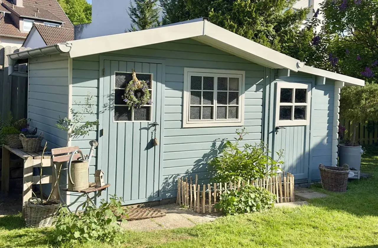 Ein hellblaues Gartenhaus mit wei&szlig;en Fensterrahmen und Dach&uuml;berstand. Davor ein kleiner Busch, daneben ein Stuhl mit Gie&szlig;kanne. Tolle gartenhaus farben ideen f&uuml;r ein charmantes Ambiente.