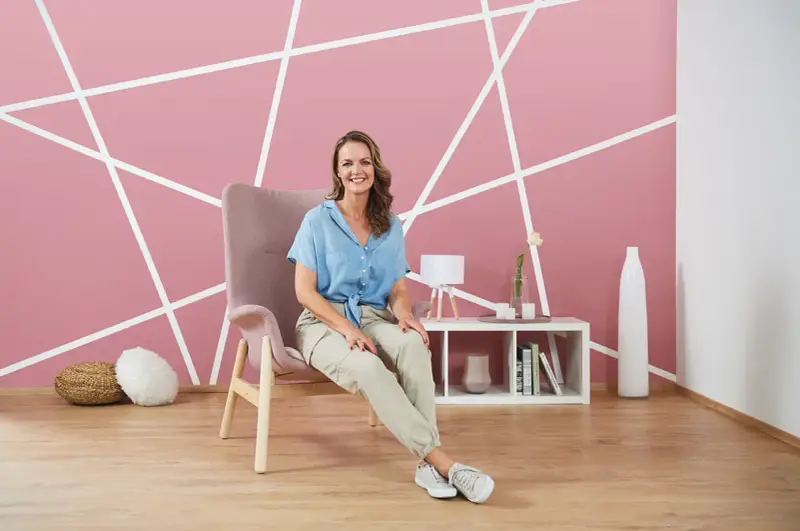 Frau sitzt vor einer rosa Wand mit weißem geometrischem Muster. Tolle Ideen zum Wand streichen und Muster abkleben!